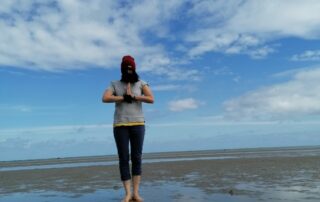 Foto Frau, barfuß am Strand in meditativer Haltung die Hände vor der Brust zusammengelegt, hinter ihr der blaue Himmel